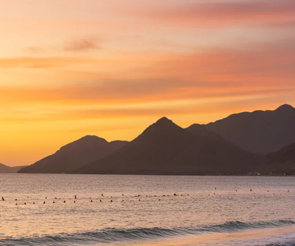 Beautiful sunset over Khor Fakkan Beach with golden sky reflecting on calm turquoise waters and Hajar Mountains silhouette