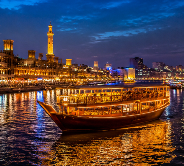 Dubai Creek Dhow Cruise – Traditional Heritage Experience Traditional wooden dhow sailing along Dubai Creek with illuminated heritage buildings of Deira and Bur Dubai at night